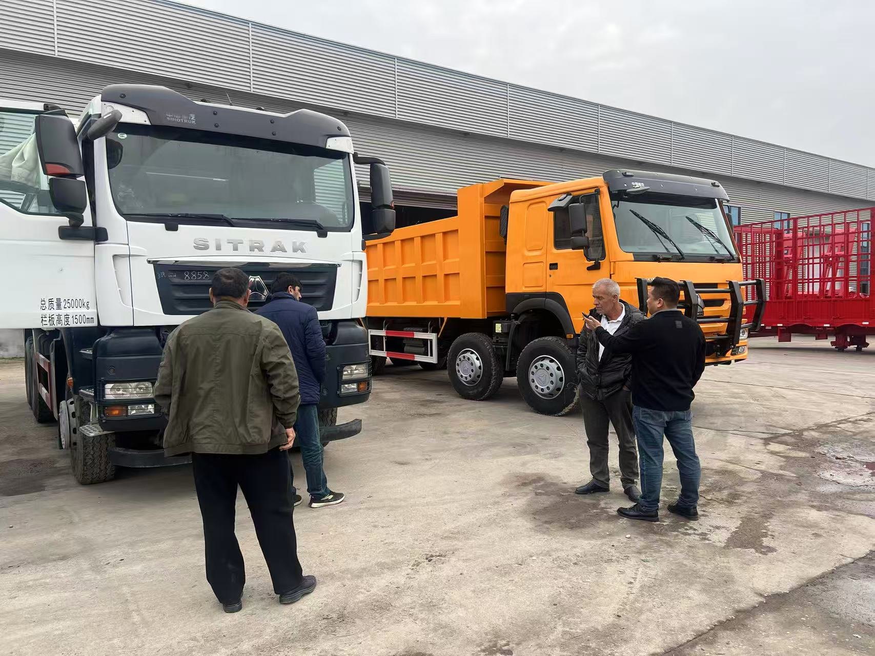 Visitors viewing dump trucks and trailers at a vehicle yard.